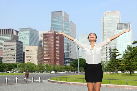 Happy free businesswoman cheering open arms in achievement and success, Japanese city woman carefree in front of Tokyo downtown skyline from the outer imperial palace gardens.