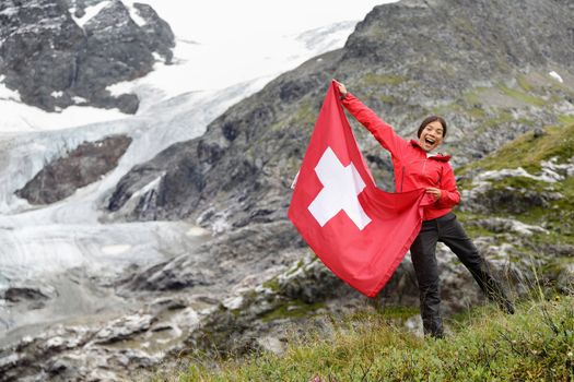 Switzerland hiker hiking cheering showing Swiss flag jumping in front of glacier. Happy Asian woman holding big red flag in nature.