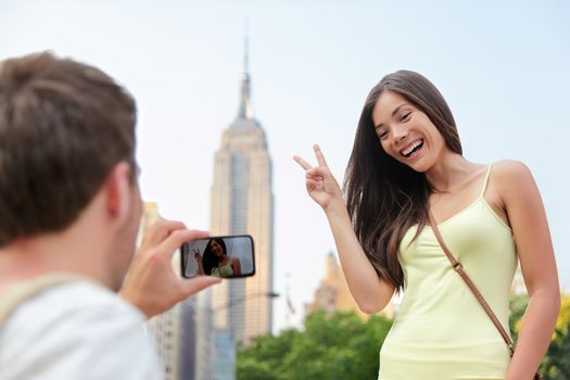 NYC asian chinese tourist girl posing at Empire State Building doing the v hand sign. Young couple of tourists taking pictures with smartphone in New York City in front of famous landmark building.