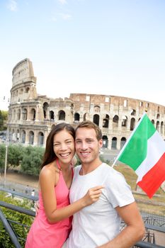Happy travel - couple tourists in front of Coliseum, Rome, Italy. Cheerful young Asian and Caucasian adults posing with Italian flag during their European holiday vacations in Europe.