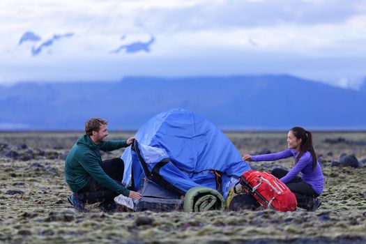 Tent - people pitching tent on Iceland at dusk. Couple setting up camp for night after hiking in the wild Icelandic nature landscape. Multicultural Asian woman and Caucasian man healthy lifestyle.