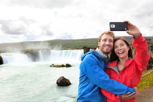 Selfie couple taking smartphone picture of waterfall Godafoss outdoors on Iceland. Couple visiting famous tourist attractions and landmarks in Icelandic nature landscape. Mixed race couple having fun.