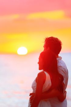 Couple watching romantic sunset during beach holidays. Newlyweds in a happy relationship at sunset during travel holidays vacation getaway. Interracial couple, Asian woman, Caucasian man.