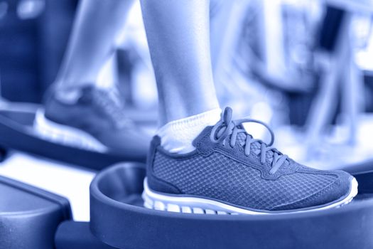 Cardio exercise - Elliptical workout machine in gym. Closeup of female feet using equipment at the fitness center for working out the legs. Sports and recreation concept in Blue monochrome filter.