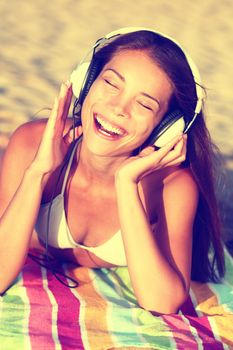 Woman listening to music with headphones at beach. Young Asian girl relaxing during summer holidays lying down with towel on golden sand enjoying her vacations singing happy.