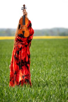 Young woman in red dress with violin in green meadow.