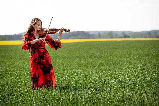 Young woman in red dress playing violin in green meadow.