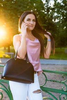 Portrait of a young woman with a handbag and a phone in the park at sunset.