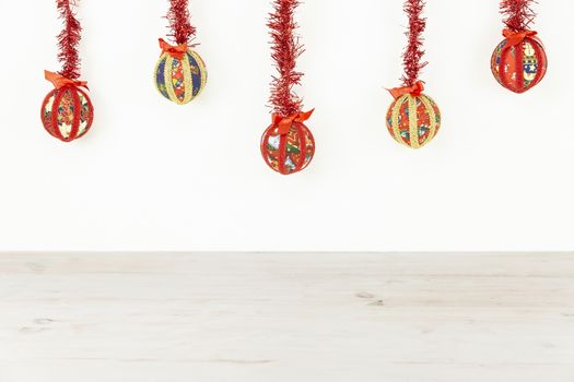 Christmas copy space: five Christmas baubles made by hand with the decoupage technique hung from top with a red shiny garland on white background and light wooden base on bottom