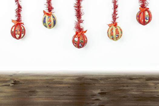 Christmas copy space: five Christmas baubles made by hand with the decoupage technique hung from top with a red shiny garland on white background and dark wooden base on bottom