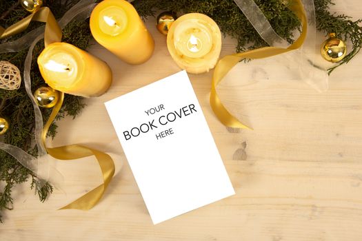 Christmas flat lay for a book presentation: book with blank cover settled with three lit candles , white organza and gold satin ribbons, gold baubles on light wooden table and dark wood background