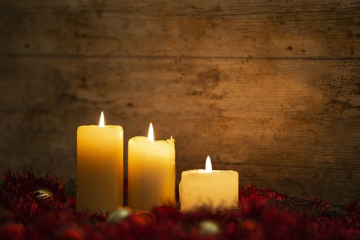 The warmth of the Christmas concept: three candles lit on a light wooden table and a rustic setting with pine branches, red decoration and gold and red bright baubles with bokeh effect