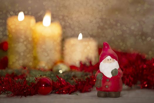 The warmth of the Christmas concept: a Santa Claus statuette in foreground in a rustic setting with with three candles lit in bokeh effect, gold and red bright baubles, snow effect