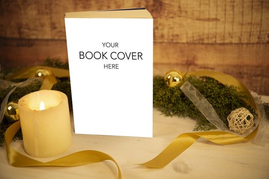 Christmas setting for a book presentation: book with blank cover settled with three lit candles , white organza and gold satin ribbons, gold baubles on light wooden table and dark wood background