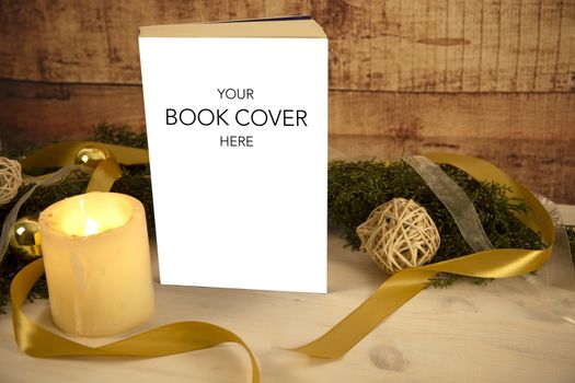 Christmas setting for a book presentation: book with blank cover settled with three lit candles , white organza and gold satin ribbons, gold baubles on light wooden table and dark wood background