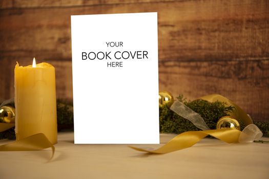 Christmas setting for a book presentation: book with blank cover settled with three lit candles , white organza and gold satin ribbons, gold baubles on light wooden table and dark wood background