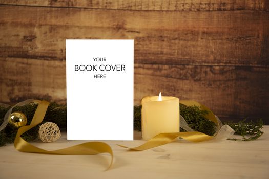 Christmas setting for a book presentation: book with blank cover settled with three lit candles , white organza and gold satin ribbons, gold baubles on light wooden table and dark wood background