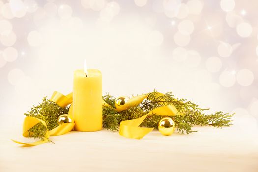 Christmas copy space with a lit yellow candle, pine branches, golden satin ribbon and gold-colored Christmas balls on a light patterned background