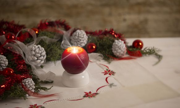 Christmas setting: a red lit candle with cross screen effect on foreground surrounded by pine branches, red baubles, red and white ribbons, white pine cones on Christmas tablecloth in bokeh effect