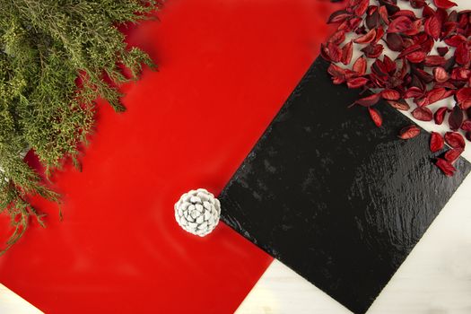 Flat lay geometric Christmas copy space with a diagonal red stripe , a black limestone plate, pine branches, white pine cone and red petals on light wooden background