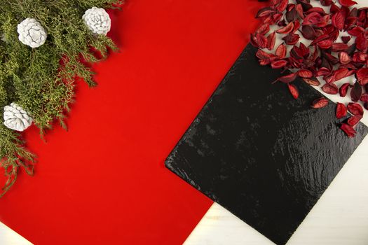 Flat lay geometric Christmas copy space with a diagonal red stripe, a black limestone plate, pine branches, white pine cones and red petals on light wooden background