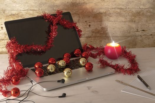 Computer job at Christmas holidays concept: an aluminum laptop open, red wreath decoration, red and gold baubles, lit candle, pen and ruler on block notes on light wooden table