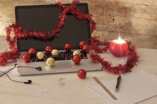Computer job at Christmas holidays concept: an aluminum laptop open, red wreath decoration, red and gold baubles, lit candle, pen and ruler on block notes on light wooden table