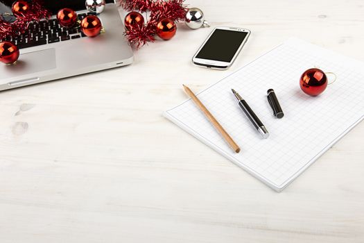 Computer job at Christmas holidays concept: light wooden table with an open aluminum laptop, red decoration, red baubles, smartphone, block notes, pencil and pen on block note