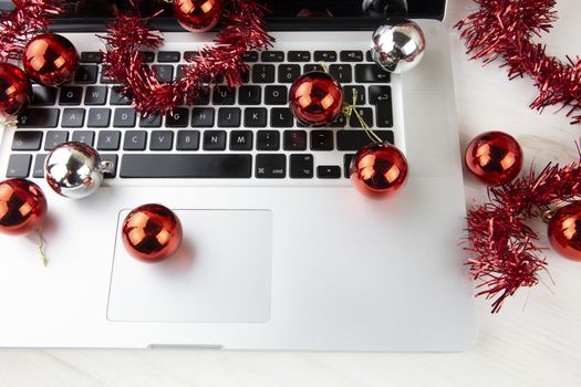 Computer job at Christmas holidays concept: close up in low angle view of an aluminum laptop open, red wreath decoration and red and silver baubles on a light wooden table