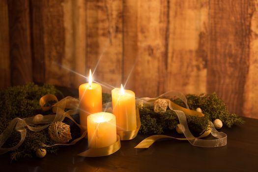 The warmth of the Christmas concept: three candles lit with cross screen stars effect on a dark wooden table with pine branches, natural pine cones with gold satin and white organza ribbons
