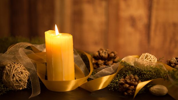 The warmth of the Christmas concept: one candle lit on a dark wooden table and a rustic wooden setting with pine branches, natural pine cones and gold satin and white organza ribbons