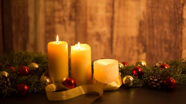 The warmth of the Christmas concept: three candles lit on a dark wooden table and a rustic setting with pine branches, natural pine cones and gold and red bright baubles with a gold satin ribbon