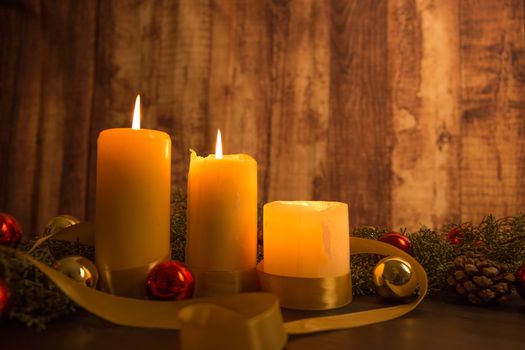 The warmth of the Christmas concept: close up of three candles lit on a dark wooden table with pine branches, natural pine cones and gold and red bright baubles with a gold satin ribbon