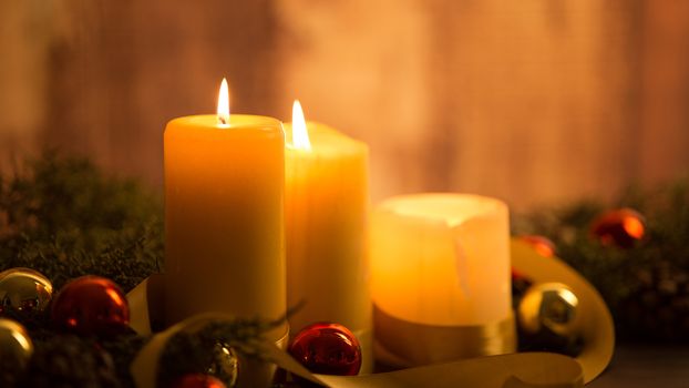 The warmth of the Christmas concept: close up of three candles lit on a dark wooden table and with pine branches, natural pine cones and gold and red bright baubles with a gold satin ribbon