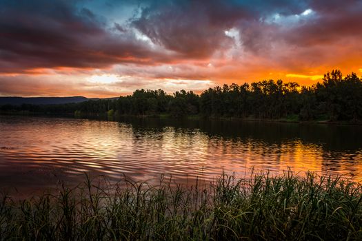 Moody stormy clouds and sunset over mountains and river with foreground grasses lining the riverbank. A tranquil place to relax, picnic, kayak or fish