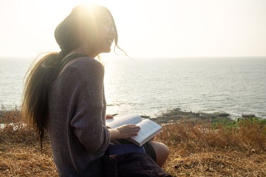 Attractive Asian woman is sitting on the edge of the mountain with a sea view, reading a book. From the back view