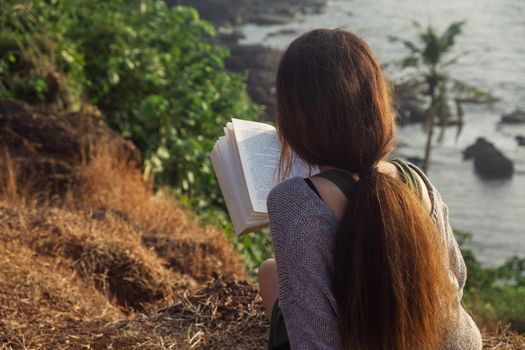 Attractive Asian woman is sitting on the edge of the mountain with a sea view, reading a book. From the back view