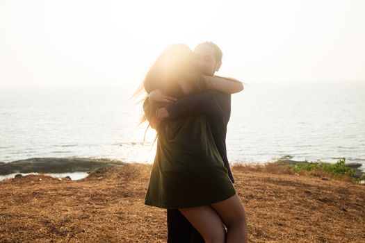 Backlit Portrait of a young romantic couple swirls in a hug against the backdrop of the sea. Anjuna Hills, Goa, India