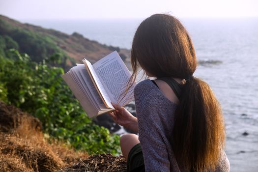 Attractive Asian woman is sitting on the edge of the mountain with a sea view, reading a book. From the back view