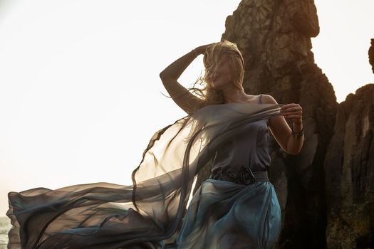 Beautiful European girl in a long dress stands on the rocks by the sea