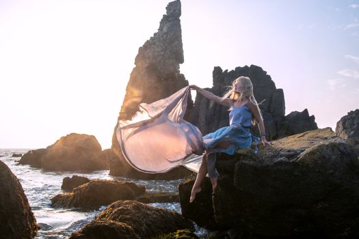 Beautiful European girl in a long dress sits on the rocks by the sea