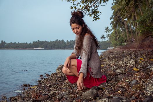 Girl with dreadlocks sits on a rocky beach and looks at the stones.