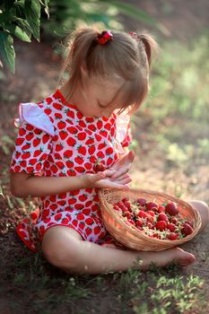 A little girl in a dress with a drawing of strawberries sits in a clearing and holds strawberry in her hands