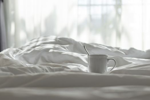 White coffee mug and spoon lay on the bed with the light from the window in the morning.