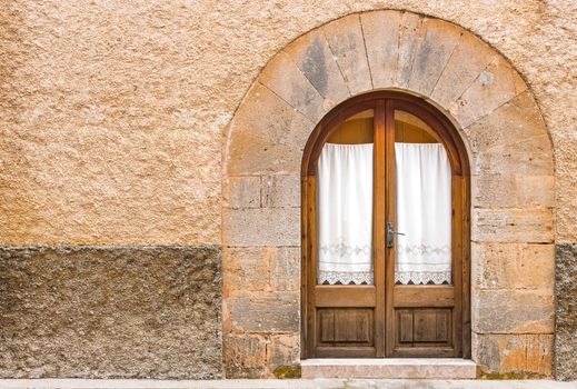 Detail view of wooden door with glass and curtain with doorway arch and stone wall of mediterranean house
