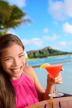 Cocktail woman drinking alcohol drinks at beach bar resort in Waikiki, Honolulu city, Oahu, Hawaii, USA. Asian girl tourist looking at camera toasting a glass of strawberry Hawaiian drink at sunset.