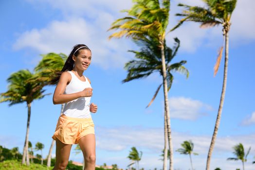 Asian runner girl jogging in tropical nature park outdoors with palm trees background during summer. Young adult sport woman running staying fit wearing orange fitness shorts and tank top.