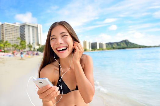 Happy beach woman listening to music on smartphone putting earphones in ears. Relaxing casual girl in bikini during summer holidays walking having fun streaming audio with wireless 4g on mobile phone.