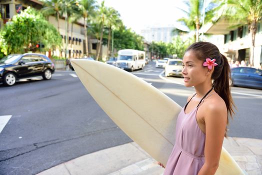 Urban surfer Asian girl holding surf board walking in city going surfing in Waikiki beach, Honolulu, Oahu, Hawaii, USA. Woman waiting for street traffic carrying surfboard and wearing Hawaiian flower.