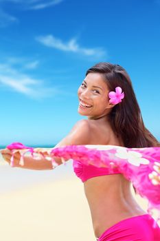Happy carefree woman in pink bikini on Hawaiian white sand beach vacation holidays holding a scarf in the wind as freedom and relaxation concept. Exotic Asian girl wearing a Hawaii flower in the hair.